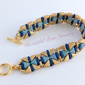 Puede incluir: Pulsera de cuentas doradas y azules con cierre de palanca y cierre de anillo. La pulsera presenta un patrón geométrico de triángulos y cuadrados. "Beautiful Rain Jewelry" está escrito en la pulsera.
