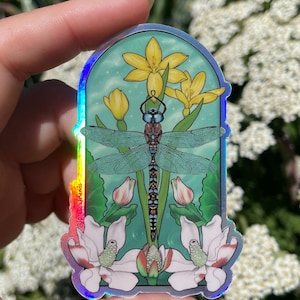 May include: An arched holographic sticker featuring a central blue dragonfly with detailed wings, positioned above white magnolia flowers and below yellow lily-like blossoms against a light green background. The text @forestxfriends is visible.