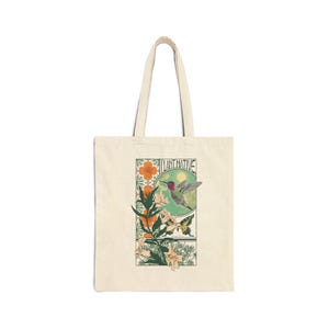 May include: Cream-colored tote bag with a rectangular graphic featuring a hummingbird, flowers, and butterflies. The graphic includes the text "PLANT NATIVE" in a decorative font. The bag has two handles for carrying.