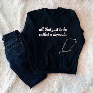 May include: A black long-sleeve crewneck sweatshirt featuring the white text "all that just to be called a dependa" printed on the chest, accompanied by a pair of dark blue denim jeans and a delicate silver-toned chain necklace with small beads.