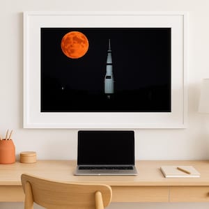 May include: Framed art print featuring a vibrant orange moon and a white rocket against a black night sky. The artwork is displayed above a wooden desk with a laptop, lamp, and decorative items. The overall theme is space and astronomy.