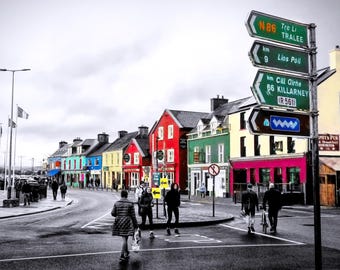 May include: A street scene in a town featuring a tall metal signpost with green directional signs pointing to Tralee, Lios Poll, and Killarney. A row of colorful buildings in red, green, and yellow lines the road, with pedestrians walking.