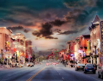May include: A street view of a city downtown area at dusk with a dramatic cloudy sky. The road is lined with multi-story brick buildings featuring illuminated neon signs, including Ole Red and Downtown, with cars driving along the asphalt street.