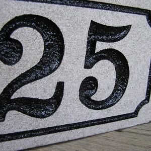May include: A white stone house number plaque with black painted numbers "25" in a decorative font.