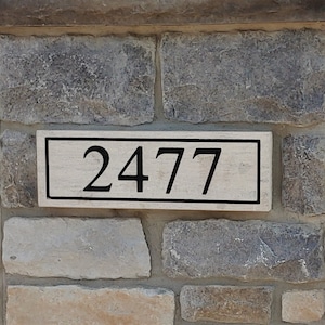May include: A rectangular address plaque with the number "2477" in black lettering. The plaque is light-colored with a black border and is mounted on a stone wall.