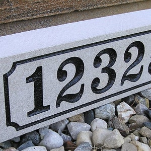 May include: A gray granite house number sign with black numbers "12325" carved into it. The sign is sitting on a bed of small gray and brown rocks.
