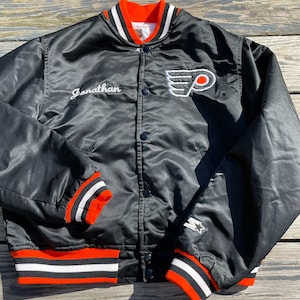 Può includere: Giacca bomber nera in satin con polsini e cintura a righe arancioni e bianche. La giacca presenta un grande logo ricamato dei Philadelphia Flyers sul petto sinistro e il nome "Jonathan" ricamato sul petto destro. La giacca ha anche una piccola stella ricamata sulla manica destra.