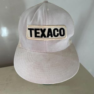 May include: A light gray baseball cap with a curved brim and mesh back. The front of the cap features a rectangular patch with the word "TEXACO" in black block letters.