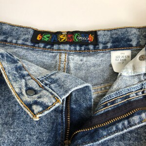 Puede incluir: Un par de pantalones vaqueros de mezclilla azul claro con cierre de cremallera y bot&oacute;n. Los vaqueros tienen un aspecto descolorido y desgastado. La cintura tiene una etiqueta negra con el texto "6*SAS*CO" en letras de colores. Una etiqueta blanca con el texto "STYLE 122 J6 578 SJ 1805 SIZE 7" est&aacute; unida a la cintura.