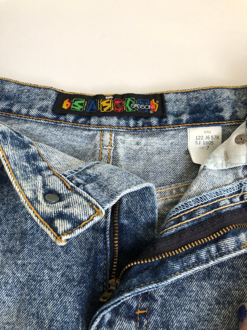 Puede incluir: Un par de pantalones vaqueros de mezclilla azul claro con cierre de cremallera y bot&oacute;n. Los vaqueros tienen un aspecto descolorido y desgastado. La cintura tiene una etiqueta negra con el texto "6*SAS*CO" en letras de colores. Una etiqueta blanca con el texto "STYLE 122 J6 578 SJ 1805 SIZE 7" est&aacute; unida a la cintura.