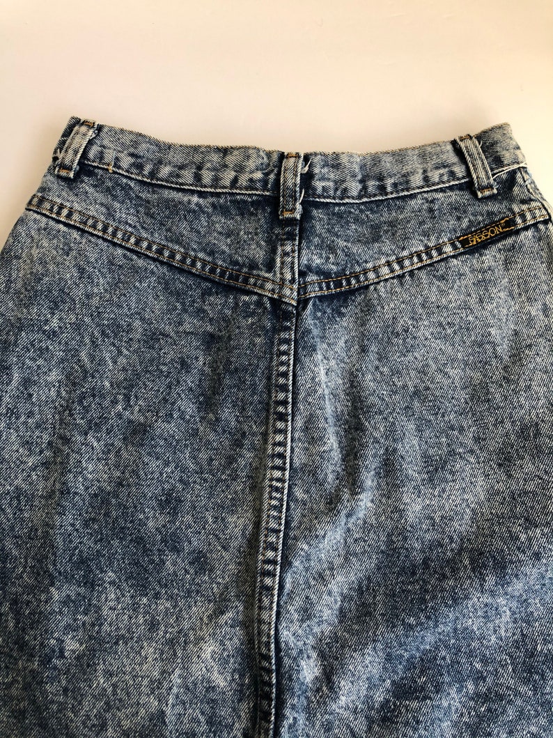 Puede incluir: Un par de jeans de mezclilla de lavado &aacute;cido de cintura alta con una etiqueta en el bolsillo trasero que dice "Guess".