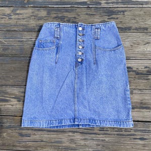 May include: A light blue denim skirt with a high waist and a button-up front. The skirt has two front pockets and two back pockets. The denim has a faded, stonewashed appearance.