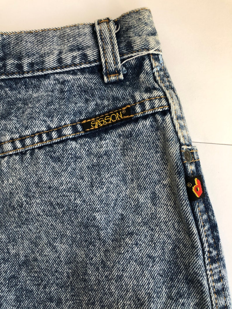 Puede incluir: Primer plano de un par de jeans de mezclilla azul claro lavados con &aacute;cido con un parche de cuero marr&oacute;n que dice "G-Star RAW" en el bolsillo trasero. Los jeans tienen un aspecto descolorido y un acabado desgastado.