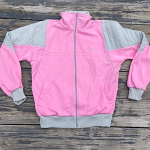 Könnte beinhalten: Eine rosa-graue Trainingsjacke mit Reißverschluss. Die Jacke hat einen hohen Kragen, grau gesteppte Schulterpartien und grau gerippte Bündchen und Saum. Ein kleines Logo ist auf der Brust sichtbar. Die Jacke liegt flach auf einer Holzoberfläche.