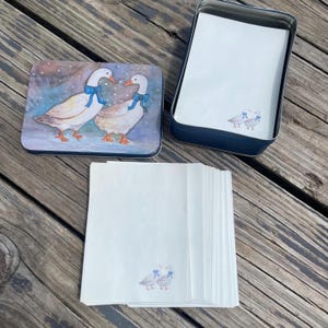May include: A rectangular metal storage tin with a lid featuring two white geese wearing blue bows, accompanied by a stack of white stationery sheets. Each sheet of paper is decorated with a small illustration of two geese with blue bows.