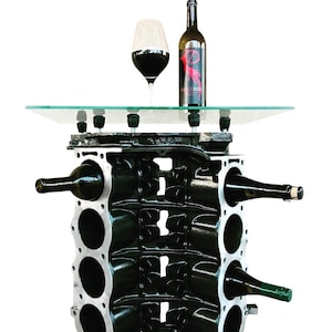 May include: A black metal wine rack made from a repurposed engine block. The rack has six slots for bottles and a glass top. A bottle of wine and a glass of red wine are on the top.