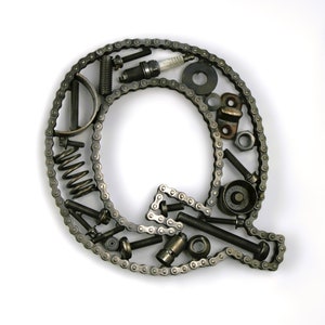 May include: The letter Q is formed using a chain and various metal parts, including nuts, bolts, springs, and a spark plug. The letter is on a white background.