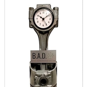 May include: A unique clock made from automotive parts. The clock face is round with black hands and white numbers. The clock is mounted on a metal connecting rod and piston. A metal plate below reads "B.A.D. BEN'S AUTOMOTIVE DECOR."