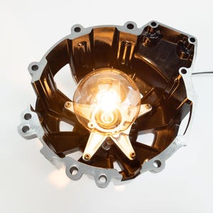 May include: A unique, industrial-style lamp featuring a repurposed metal engine part. The lamp has a warm, glowing lightbulb at its center, held in place by a star-shaped fixture. The metal casing is dark with silver edges.