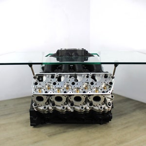 May include: A glass-topped coffee table with a car engine base. The engine is made of metal and has a silver and black finish. The glass top is clear and has a beveled edge.