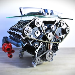 May include: A repurposed car engine transformed into a unique coffee table. The engine block is painted black with silver accents. The table features a glass top and a red water pump. The engine is mounted on wheels.