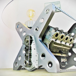 May include: A silver metal table with a glass top. The table is made from a repurposed car engine. The engine block is visible and has the word "BRODIX" on it.