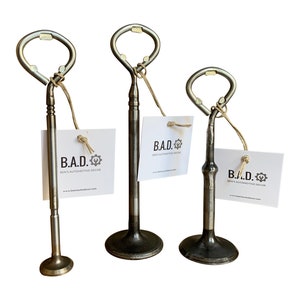 May include: Three metal bottle openers shaped like car engine valves. Each opener has a brown string attached to the top. The openers are on a white background. The text on the cards reads "B.A.D. Ben's Automotive Decor"