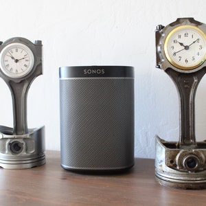 Two car piston clocks beside a speaker on a table. Left clock is finished in gunmetal gray with a silver clock ring, and the right clock has a patina finish with a gold clock ring.