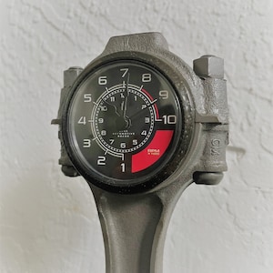 May include: A black and red automotive gauge with a white face and black numbers. The gauge reads RPM x 1000. The gauge is mounted on a gray metal rod.