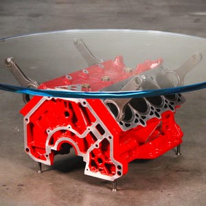 May include: A coffee table featuring a large, circular clear glass top supported by a bright red painted automotive engine block base. The base includes metallic connecting rods extending upward to hold the glass, with small metal feet at the bottom.