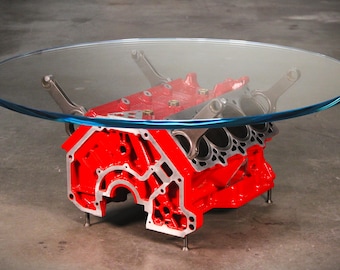 May include: A coffee table featuring a large, circular clear glass top supported by a bright red painted automotive engine block base. The base includes metallic connecting rods extending upward to hold the glass, with small metal feet at the bottom.