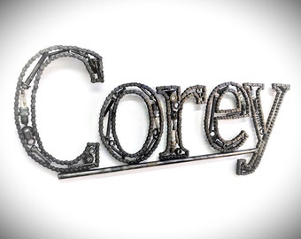 May include: The name "Corey" is spelled out using a variety of metal pieces, including chains, bolts, and nuts. The letters are connected by a metal rod.
