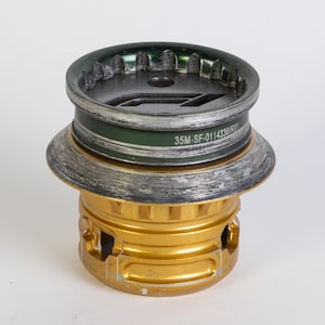 May include: A metallic gold-colored mechanical part with a dark green and silver top. The top has a series of teeth-like edges. The text "35M-SF-0114336/R0" is printed on the green band.