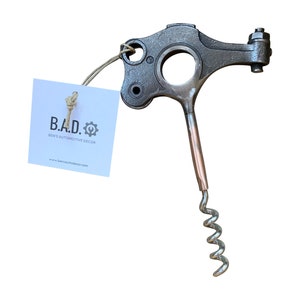 May include: A silver metal bottle opener shaped like a car part with a corkscrew attached. The bottle opener has a tag attached that says "B.A.D. Ben's Automotive Decor www.bensautodecor.com"