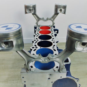 May include: A unique table crafted from automotive engine components. The table features a blue and silver engine block with red and black accents. The pistons are topped with a blue and white logo. A creative piece for car enthusiasts.