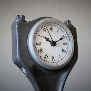 Close-up view of a clock made out of a car engine's piston, finished in gunmetal gray with a silver clock ring.