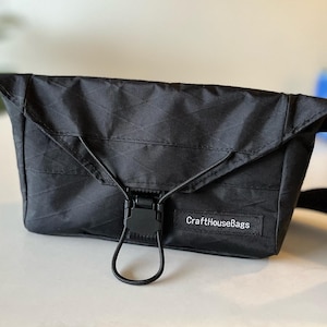 May include: Black crossbody bag with a geometric pattern and a buckle closure. The bag has a flap closure and a shoulder strap. The text "CraftHouseBags" is visible on the front of the bag.
