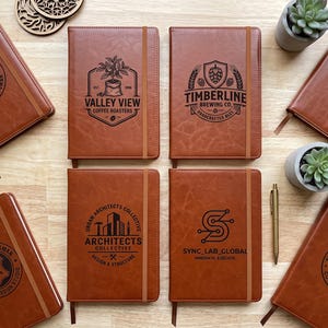 May include: A collection of eight rectangular brown notebooks with elastic closures, each featuring a unique black laser-engraved logo. The notebooks display various designs including Valley View Coffee Roasters, Timberline Brewing Co, and Urban Architects Collective.