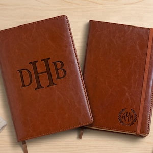 May include: Two brown faux leather journals with stitched edges. The left journal features the engraved letters DHB in a large serif font. The right journal has an orange elastic closure and a small circular laurel wreath logo with AKM inside.