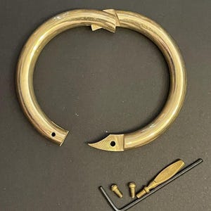 May include: A disassembled brass bracelet with a distinctive clasp design. Includes two screws, a small screwdriver, and an Allen key. The bracelet is circular, with a polished finish, against a dark background.