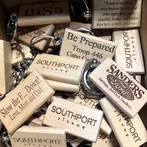 Puede incluir: Una colección de llaveros de madera con anillos de metal plateado. Los llaveros tienen varios textos grabados, incluyendo "SOUTHPORT strong", "Slow the F... Down! Love, Mom and Dad", y "Sanders Ford". Los llaveros son de color marrón claro.