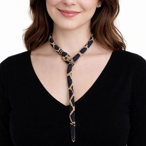 Op de afbeelding: Een kralenketting met een slangenontwerp. De ketting heeft een donkerblauw en goud patroon, met een slangenkopsluiting. De vrouw op de foto draagt een zwarte top.