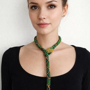 Op de afbeelding: Een kralenketting in de vorm van een slang, met groene, gele en zwarte kralen. De ketting wordt om de nek gedragen en hangt naar beneden. De achtergrond is een effen witte muur.