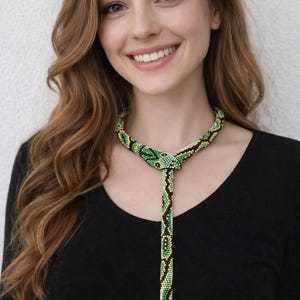 Op de afbeelding: Een kralenketting met een slangenontwerp. De ketting heeft groene, zwarte en gele kralen. De ketting wordt gedragen over een zwarte top.