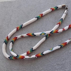 May include: A long, tubular beaded necklace featuring a white base with repeating diagonal stripes of red, orange, yellow, green, and blue beads. The necklace is finished with silver-toned metal end caps and a lobster claw clasp closure.