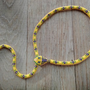 Può includere: Una collana di serpente di perline gialle con una testa di serpente di perline. Il serpente è decorato con perline blu e verdi. La collana è su una superficie di legno.