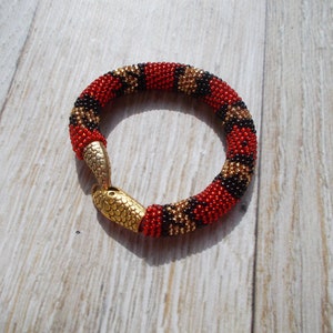 Puede incluir: Un brazalete de cuentas rojas, negras y doradas con un cierre en forma de serpiente.