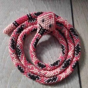Può includere: Un serpente di perline rosa, nero e argento arrotolato in un cerchio. La testa del serpente è sollevata e presenta un motivo di perline rosa e nero.