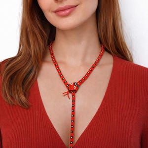 Op de afbeelding: Een rode kralenketting met een uniek ontwerp. De ketting heeft een centraal decoratief element en een lange, hangende streng. De kralen zijn rood met zwarte accenten. De ketting wordt gedragen over een rode top.