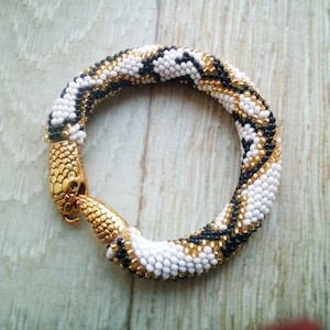 Könnte beinhalten: Ein Armband aus schwarzen, weißen und goldenen Perlen mit einem Schlangenkopfverschluss.